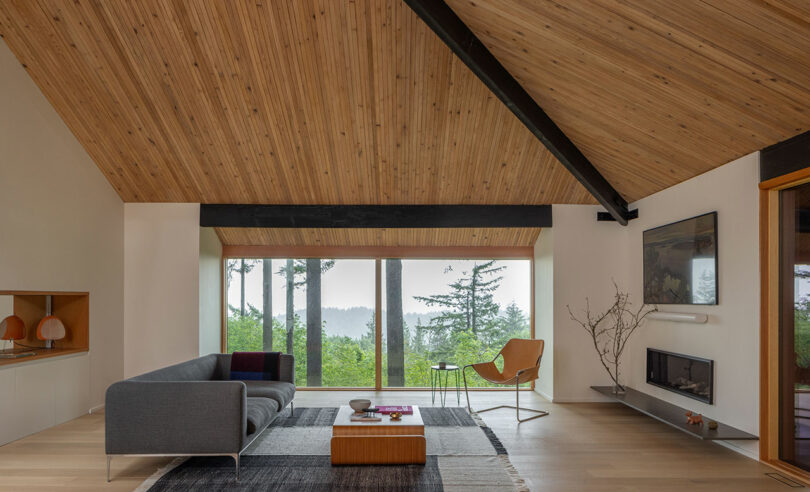 A modern living room with a wooden ceiling, large window with a forest view, gray sofa, orange chair, fireplace, and a minimalist decor.