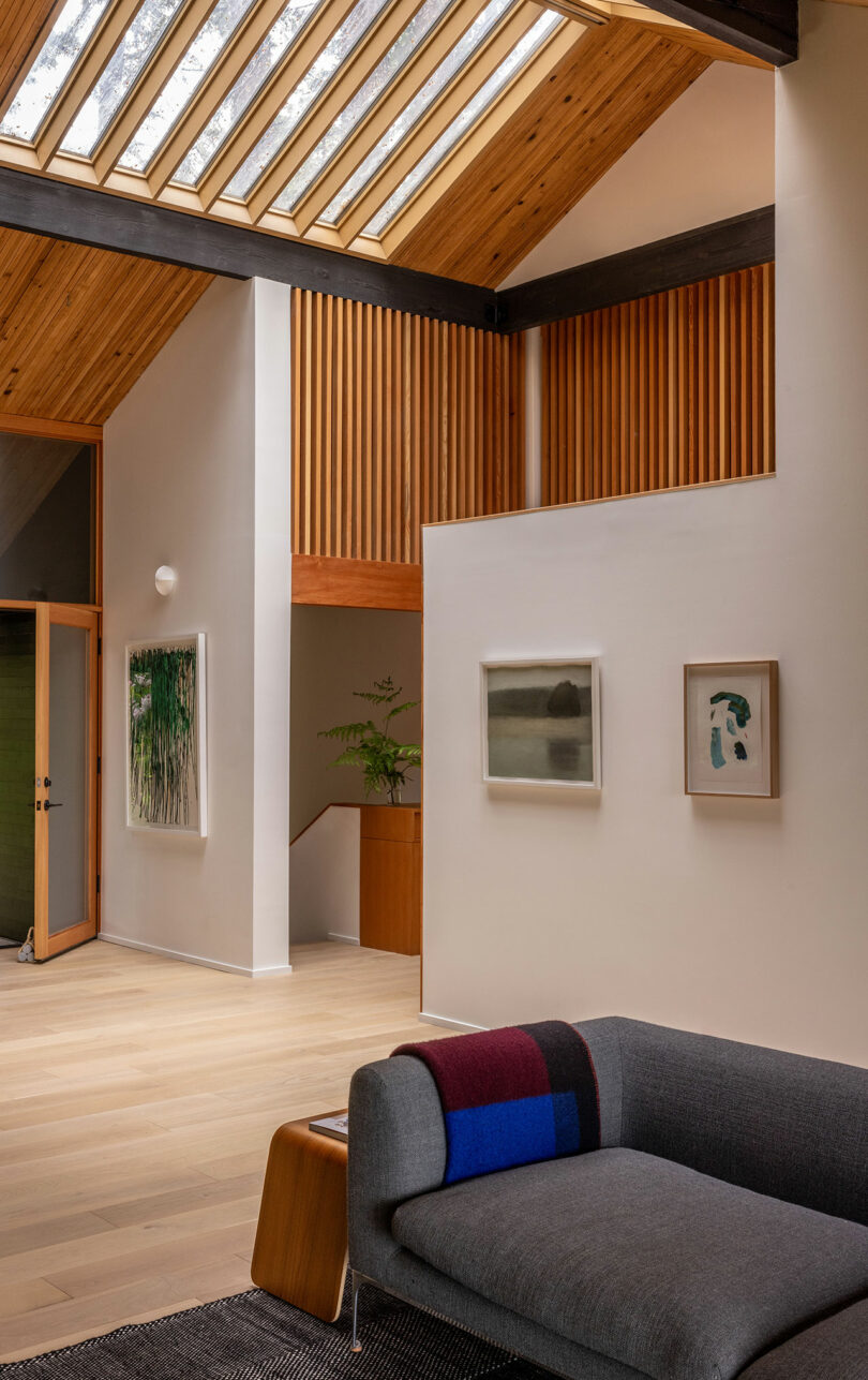 Modern living room with wooden ceiling, skylights, and minimal decor. A gray sofa with a colorful blanket faces two framed artworks on a white wall.