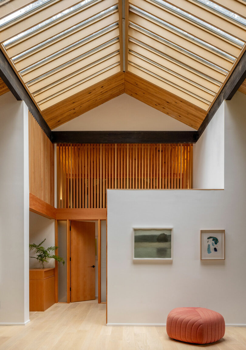 A modern interior with a high wooden-beamed ceiling, minimalist decor, two paintings on the wall, a plant, and a round pink ottoman.