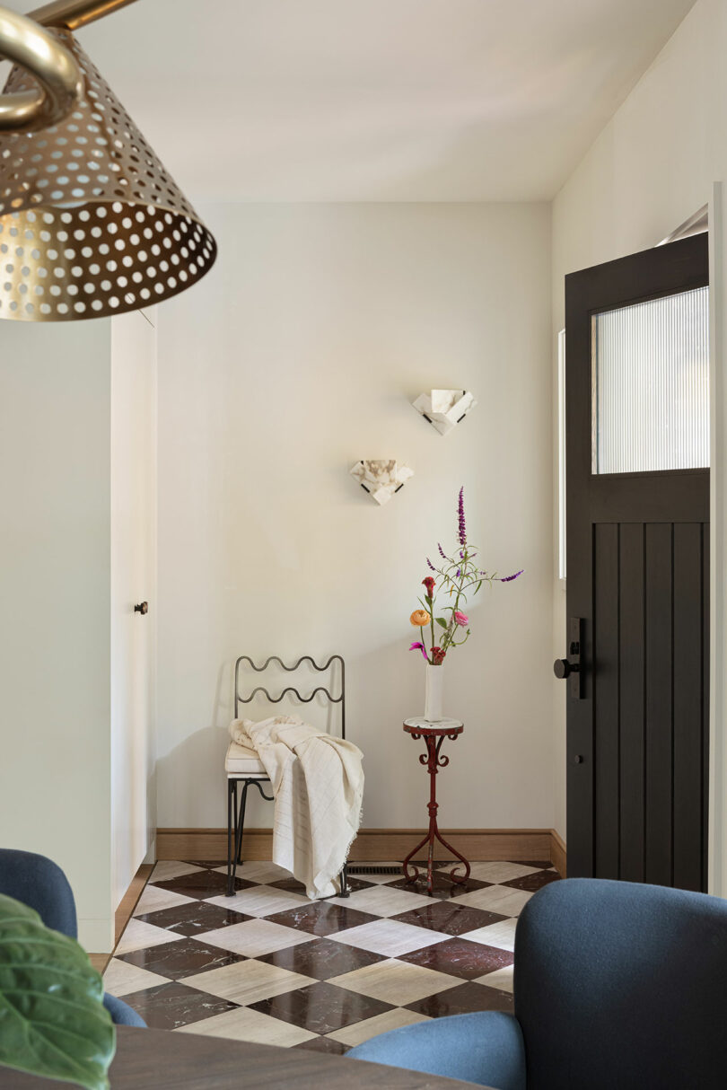 A small entryway features a black chair with a white cloth draped over it, a red side table with a vase of flowers, a black door, and a checkered floor. Two wall-mounted light fixtures are visible.