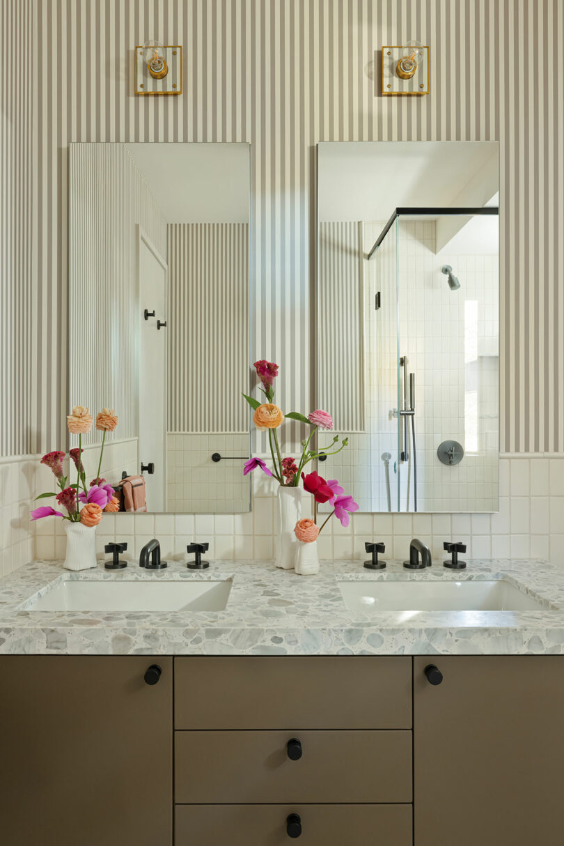 A bathroom with a double sink vanity, marble countertop, striped wallpaper, and wall-mounted mirrors. Two vases with colorful flowers are placed on the counter.