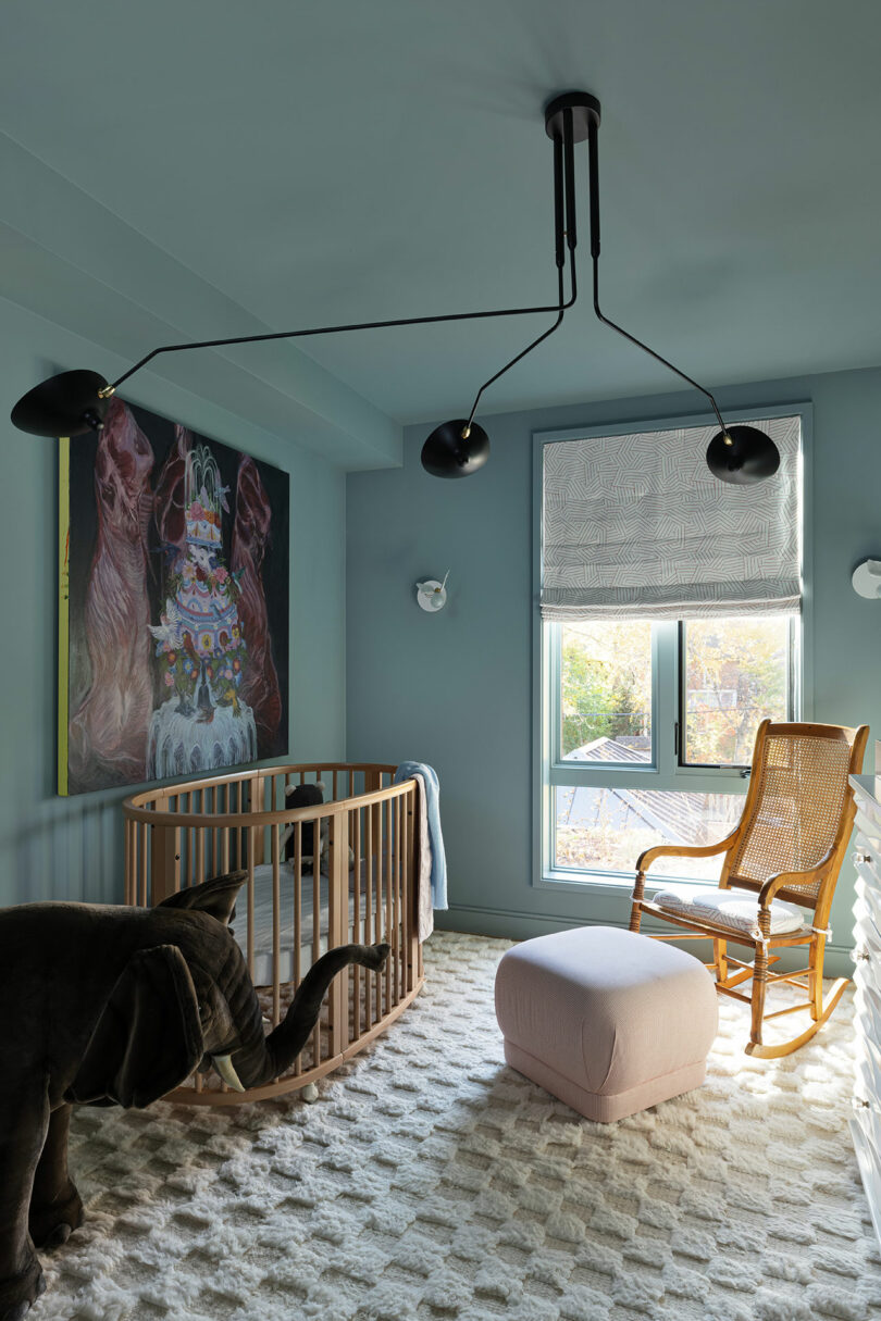 A baby room features a wooden crib, an elephant plush toy, a rocking chair, a pink ottoman, a large animal-themed artwork, and a window with a roman shade. A modern chandelier is on the ceiling.