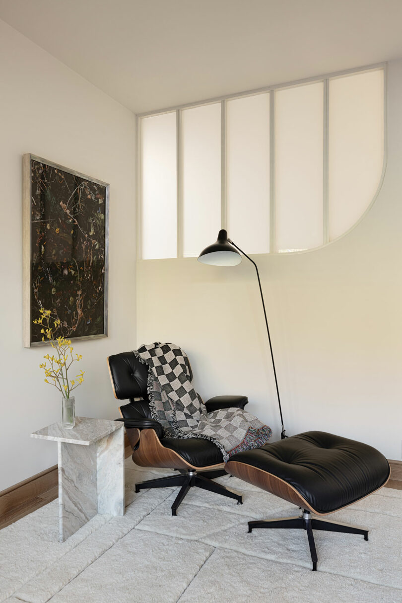 A modern lounge area features a black leather chair with matching ottoman, a tall floor lamp, a side table with a plant, and a wall painting, all set against a light-colored wall with curved detailing.