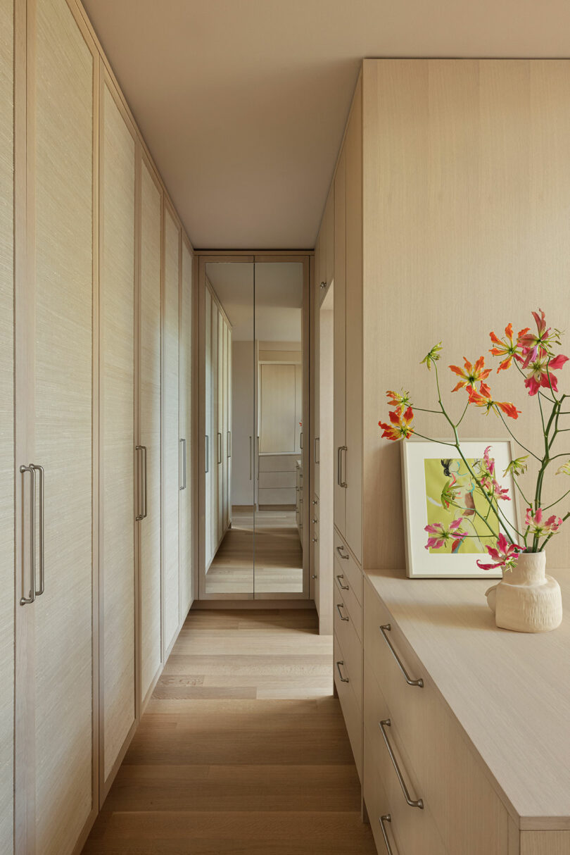 A hallway with built-in wooden wardrobes on both sides, a chest of drawers with a vase of flowers and a picture frame, and a large mirror at the end.