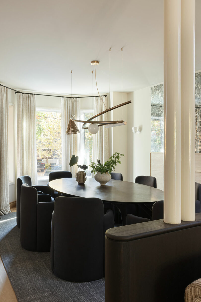 A modern dining room with a large oval table, dark chairs, and a unique overhead chandelier. The room features large windows with light curtains and a decorative plant on the table.