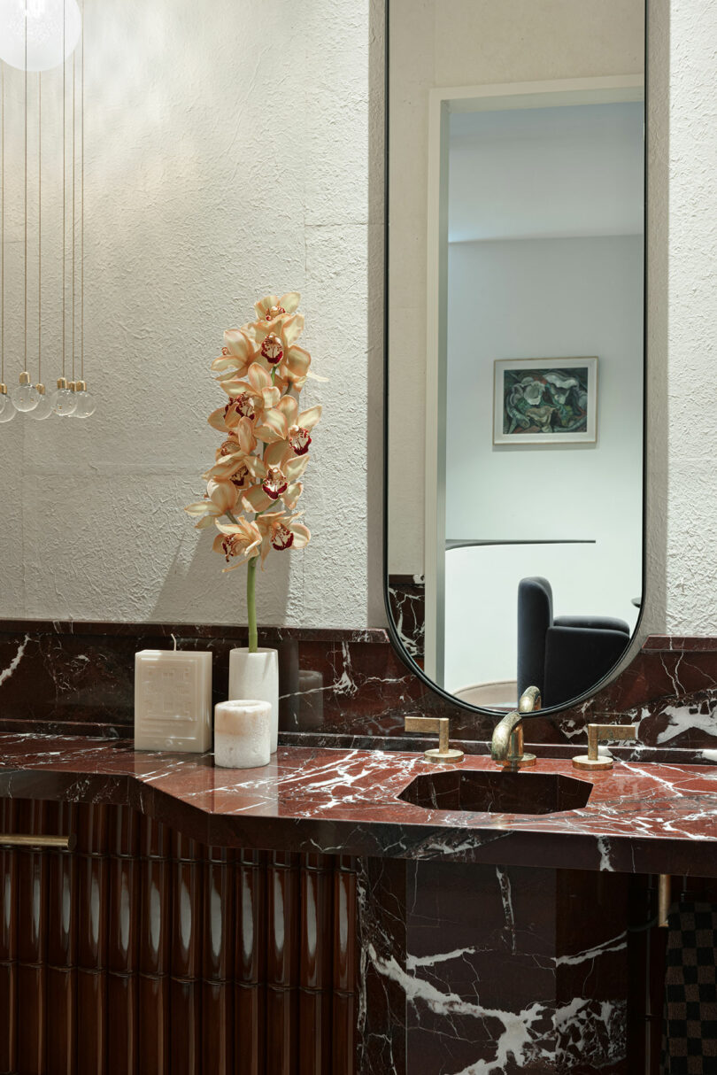 A bathroom with a large mirror, a marble countertop, a sink with a gold faucet, an orchid in a vase, and a framed picture on the wall.