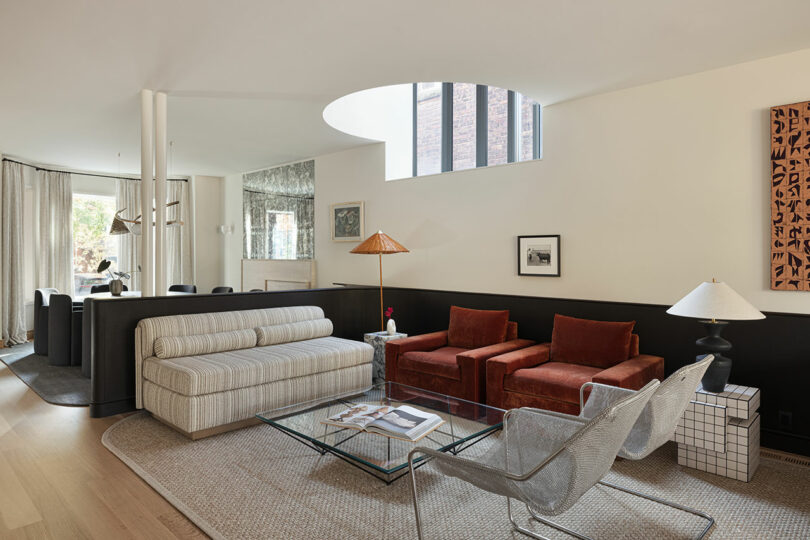 A modern living room features a round skylight, striped sofa, two red armchairs, glass coffee table, metal chairs, floor lamp with a woven shade, and wall art. Background has dining area with hanging light.