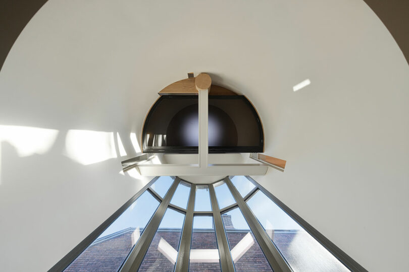 Image of a modern, semi-circular rooftop window with sunlight streaming through, showcasing a geometric ceiling design with overlapping wooden and metal elements.