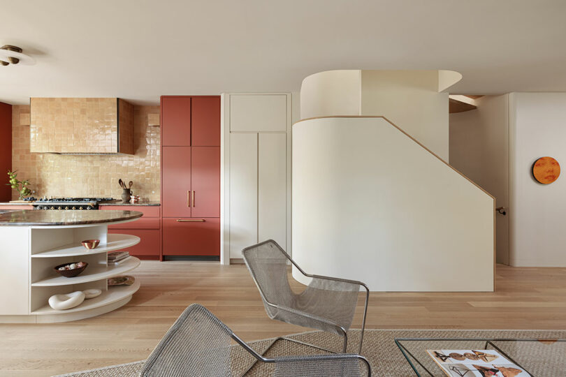 Modern kitchen and living area with light wood flooring, red cabinetry, curved white staircase, open shelving, and mesh chairs. Decor includes a wall-mounted plate and a magazine on the coffee table.