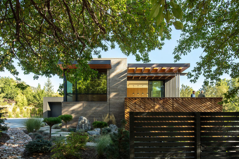 Modern house with large windows and wood accents, surrounded by trees and landscaped garden, viewed from behind a wooden fence.