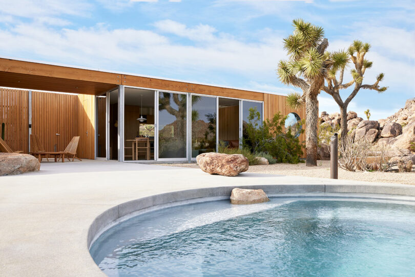 Modern house with glass walls and wooden accents, surrounded by desert landscape, featuring a small circular pool in the foreground.