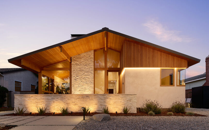 Modern house with angled roof, stone and wood facade, large windows, illuminated exterior, and landscaped front yard at dusk.