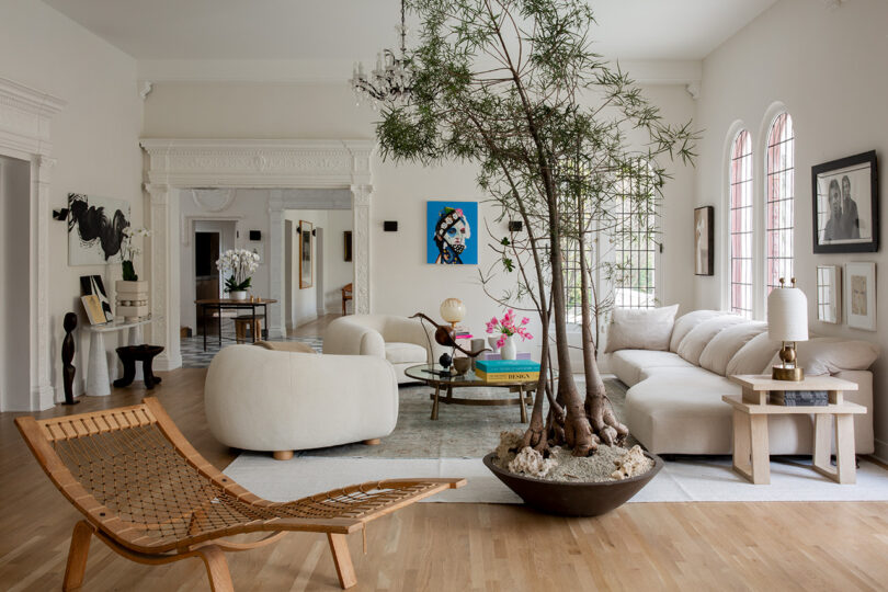 Spacious living room with modern decor, featuring a large potted tree, white sofa, wooden lounge chair, and artwork on the walls.