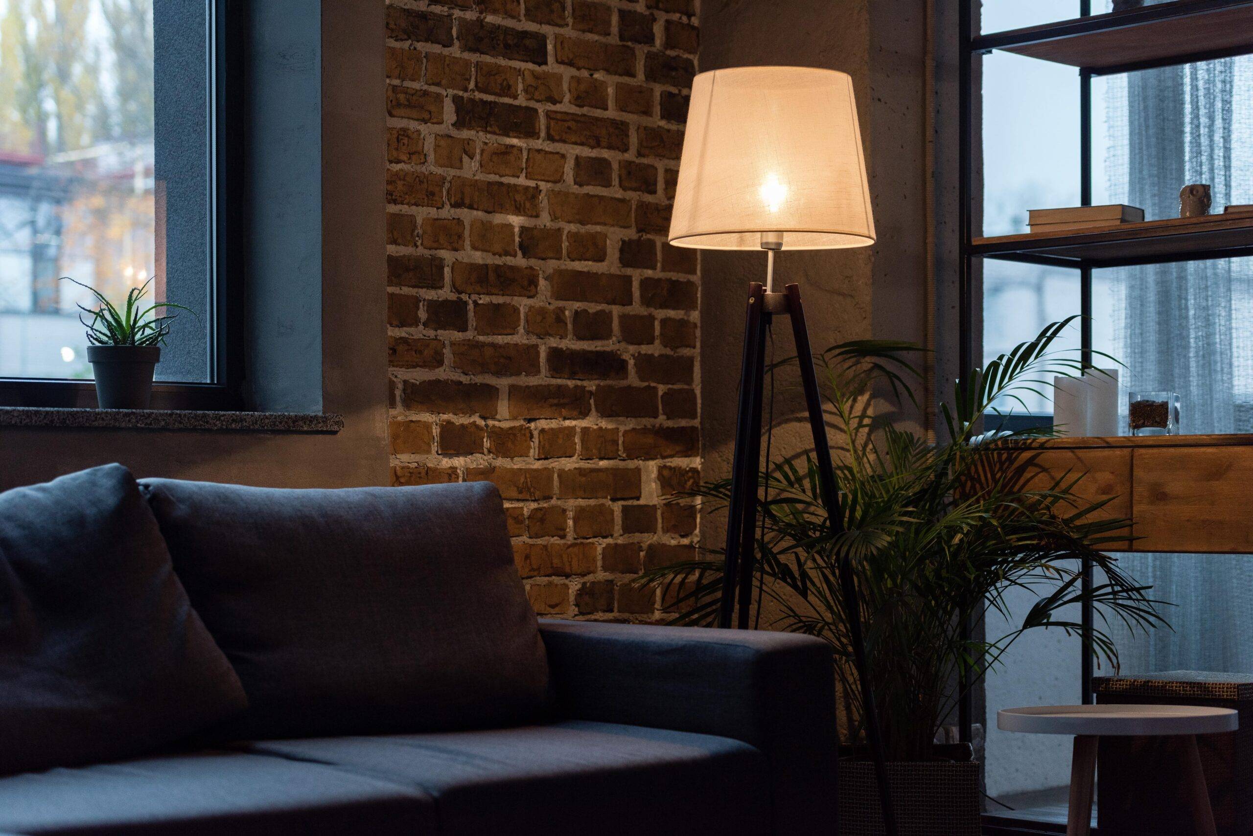 A dark yet cozy corner of an apartment, a navy couch against an exposed brick wall, a tall floor lamp beside it.