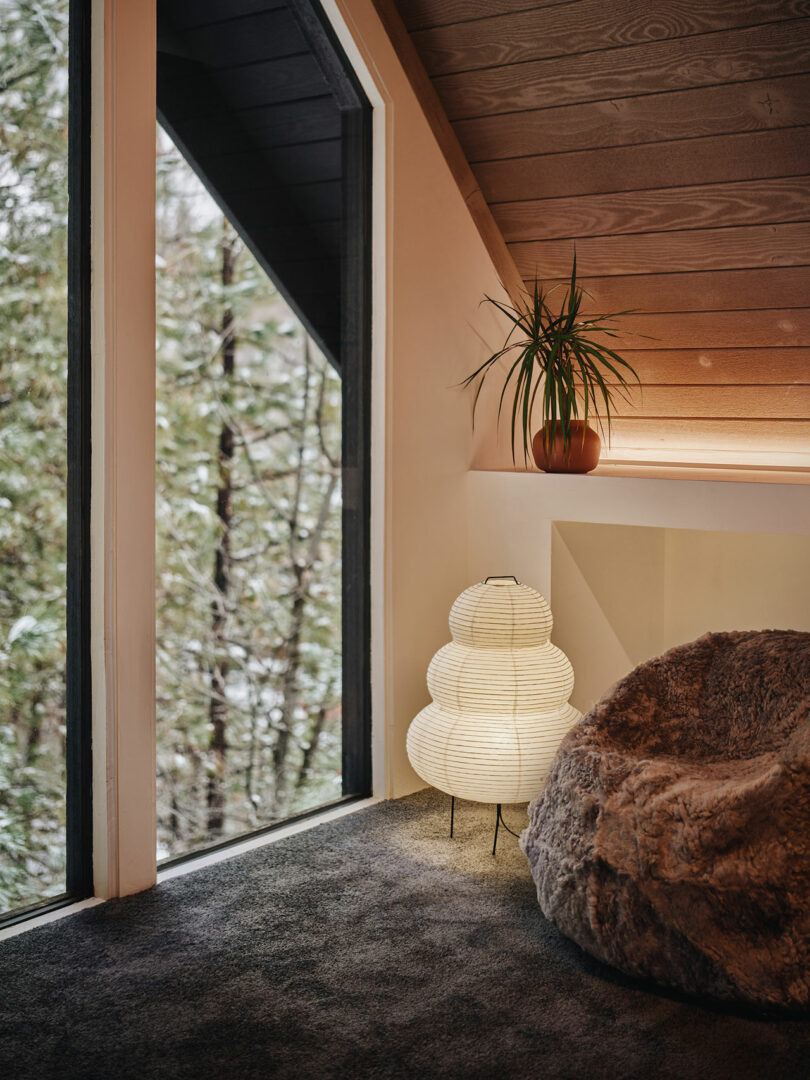 Cozy room with a large window revealing a forest view, a fluffy chair, a layered paper lamp on the floor, and a potted plant on a ledge under a slanted wooden ceiling.