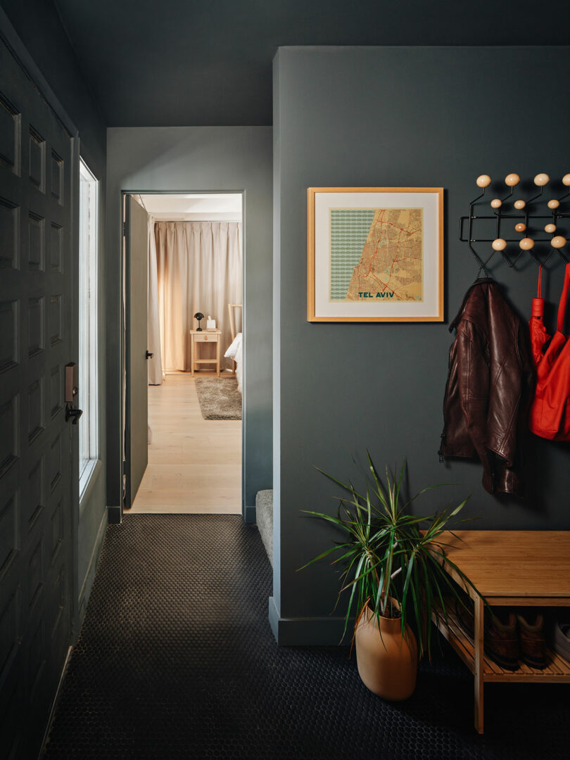 Entryway with a dark wall, coat hooks, a brown jacket, and a red bag hanging. A plant and a bench are nearby. A map of Tel Aviv is framed on the wall. A doorway leads to a bright room.