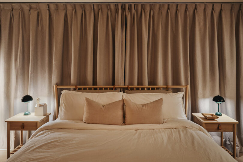 A neatly made bed with beige pillows and white bedding, flanked by two wooden nightstands with green lamps. Beige curtains cover the wall behind the bed.