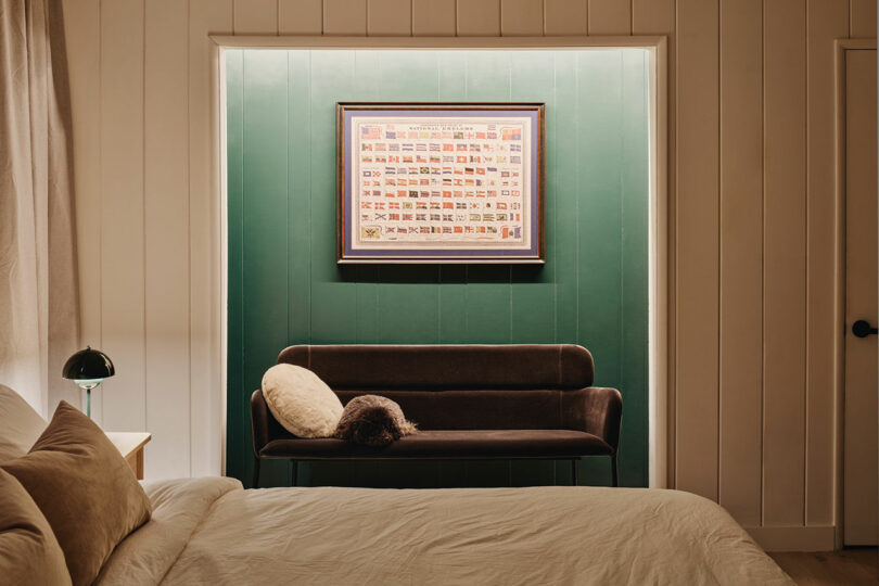 A cozy bedroom with a bed in the foreground, a dark sofa against a green wall, a framed picture above, and a small lamp on a side table.