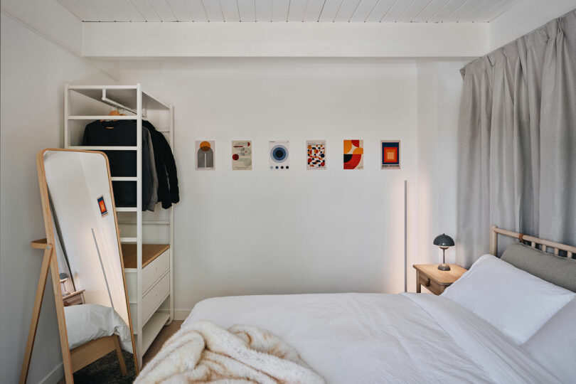 A tidy bedroom with a white bed, freestanding mirror, and wall art. A wooden nightstand holds a lamp. Artwork is reflected in the mirror.