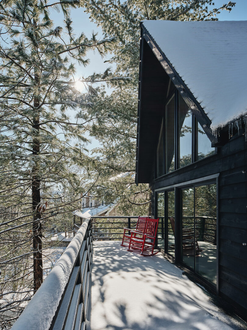 A snow-covered wooden deck with a red rocking chair. Large trees and the sun are visible in the background next to a black house with large windows.