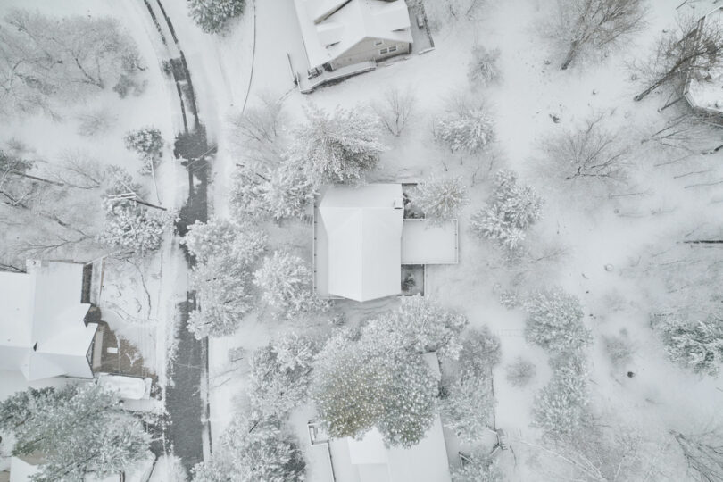 Aerial view of a snow-covered neighborhood with houses, trees, and roads blanketed in white, creating a serene winter scene.