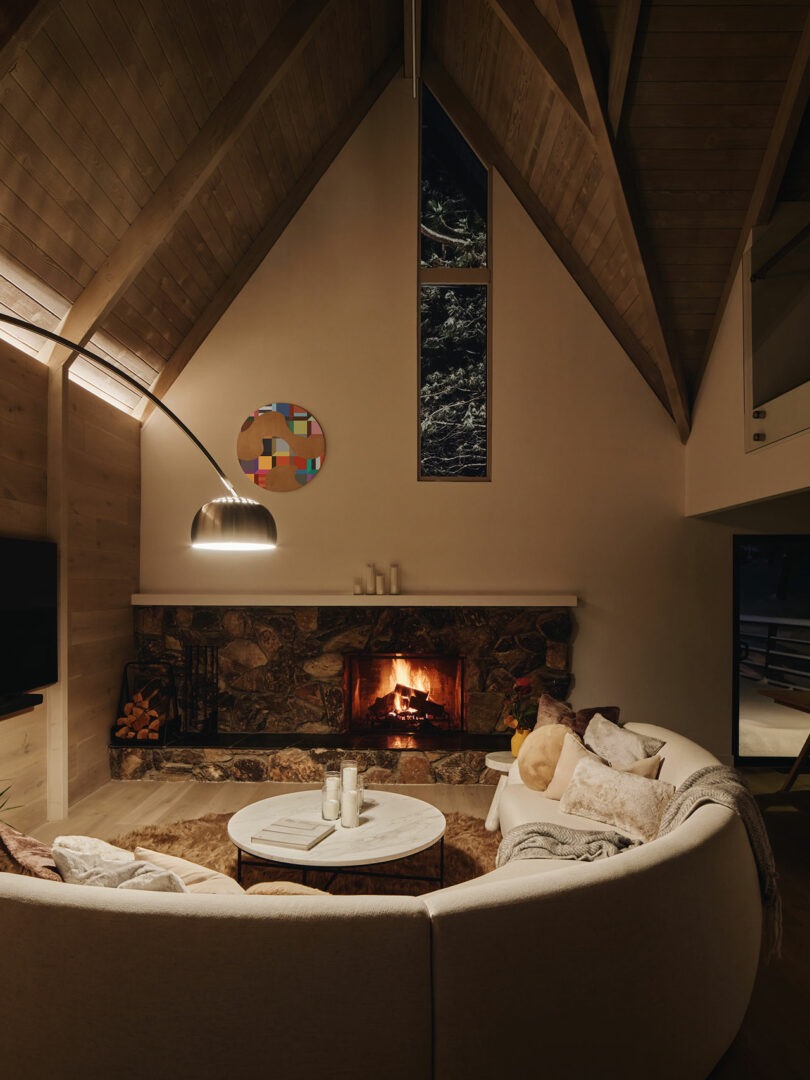 Cozy living room with a round, sectional sofa facing a stone fireplace. A tall, arched lamp hangs over the sofa, and the high ceiling features exposed wood beams.