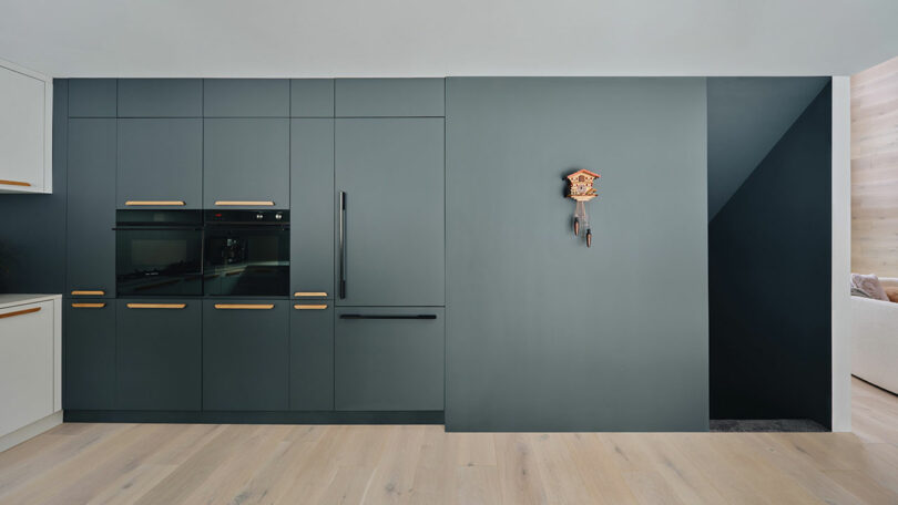 Modern kitchen with sleek gray cabinets, built-in appliances, and light wood flooring. A small decorative clock is mounted on the wall.