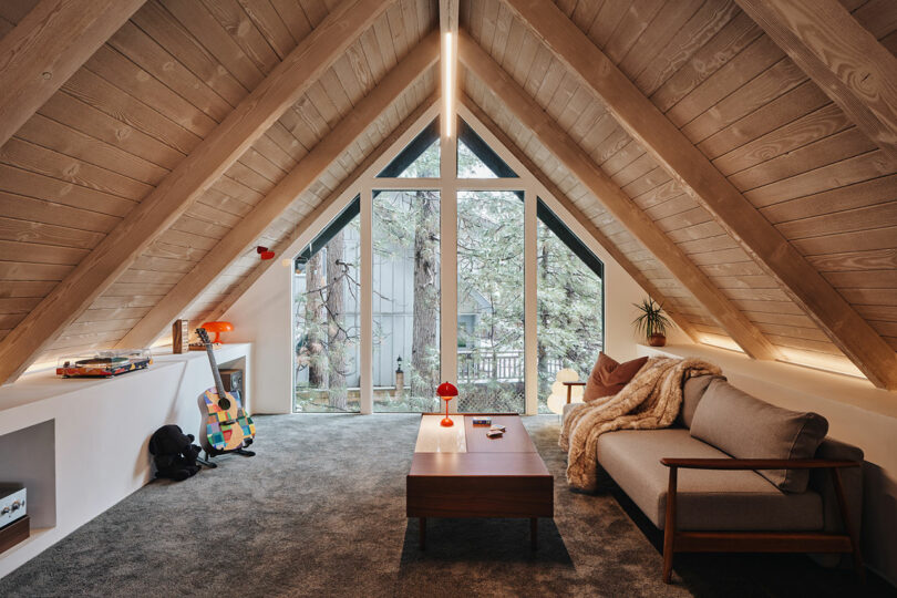 A cozy attic room with a sloped wooden ceiling features a sofa, a wooden coffee table, a guitar, and large triangular windows overlooking green trees. A small red lamp sits on the table.