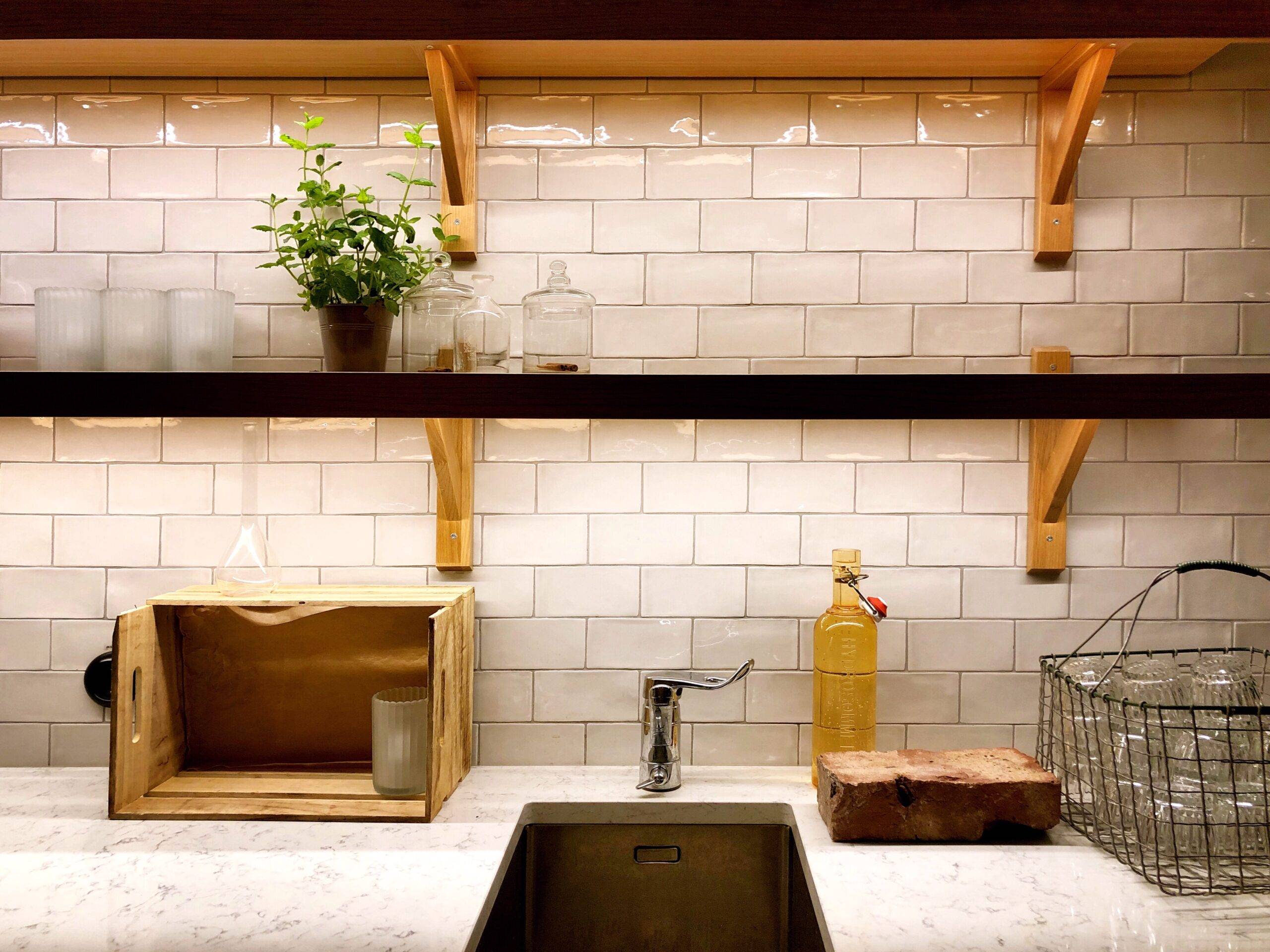 Some open shelves above a kitchen sink with a white tiled backsplash behind them.