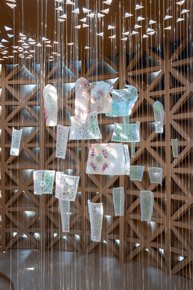 Hanging translucent sculptures with geometric patterns, suspended by strings in an artistic display against a diamond-patterned wooden wall.