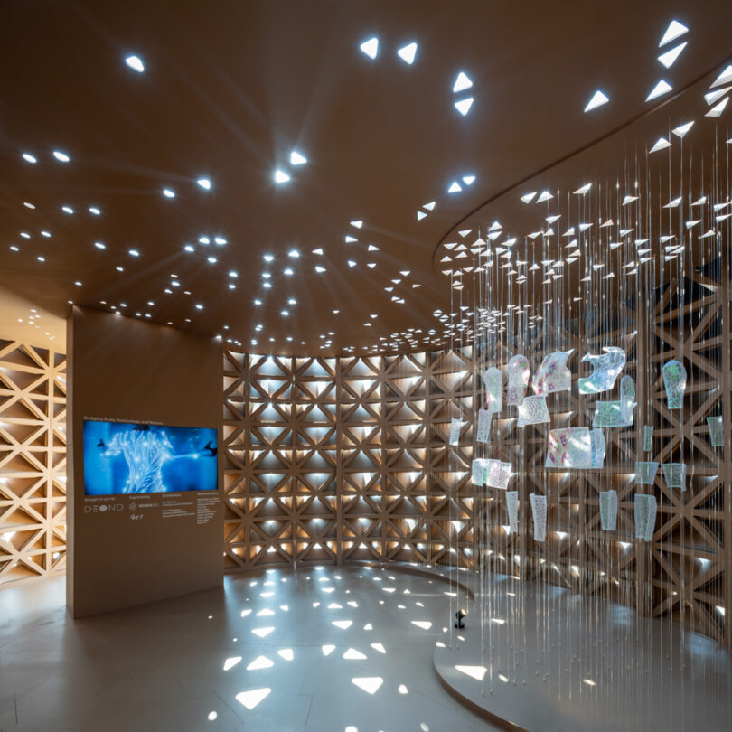 A modern exhibition space with geometric patterns on the walls, a digital display screen, and hanging translucent panels.