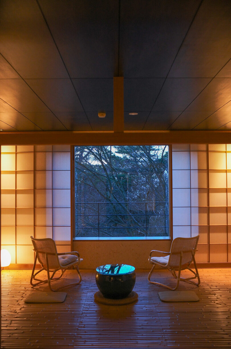 In a serene space designed by Amanda Gunawan, two chairs and a round table rest on wooden flooring, framed by a large window offering a view of the trees outside.