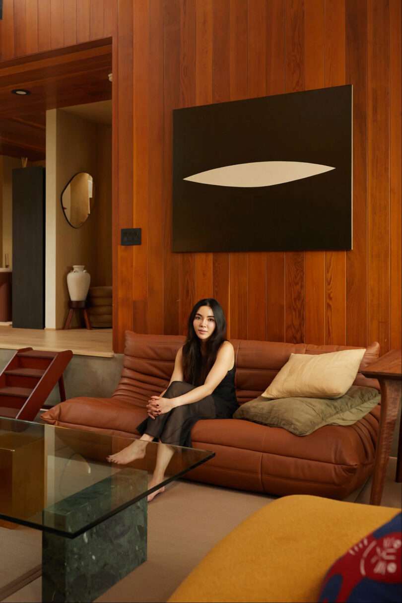 Amanda Gunawan sits on a brown leather sofa in a modern living room with wooden walls, next to a square black and white painting. A glass coffee table and decorative mirror accentuate the stylish space.