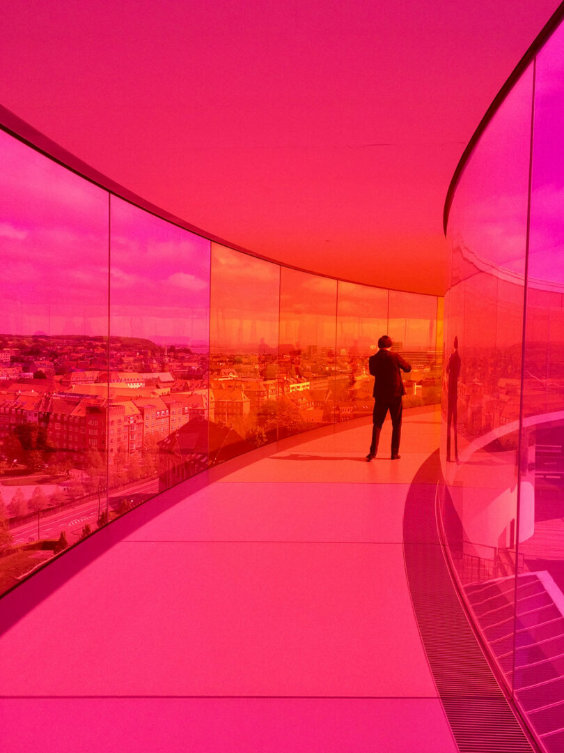 Person standing in a curved corridor with pink-tinted glass walls, overlooking a cityscape.