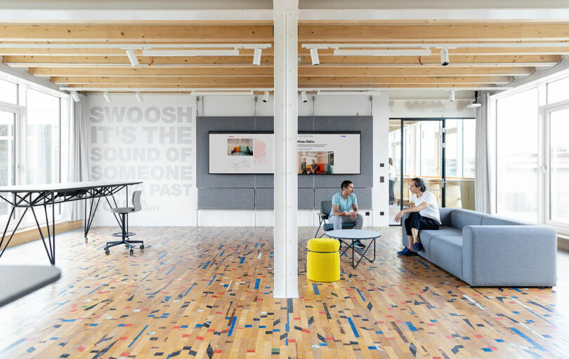 Two people sitting on a gray sofa in a modern, open office space with wooden beams, colorful floor, and wall art. A black table with a chair stands to the left.