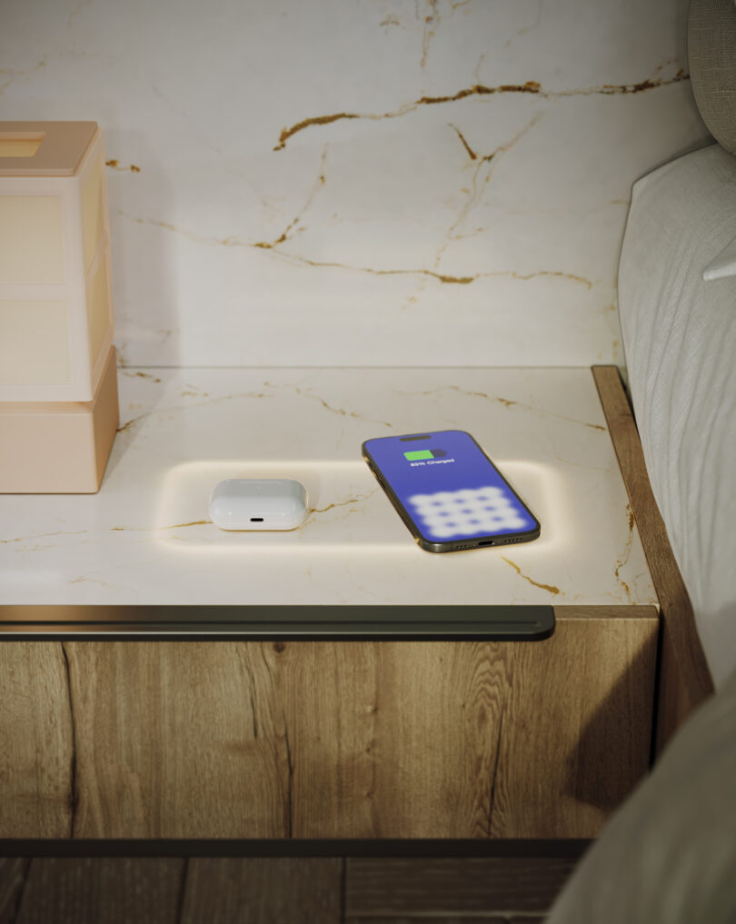 A smartphone and wireless earbuds on a wooden nightstand with a marble top. The phone screen shows a charging icon.