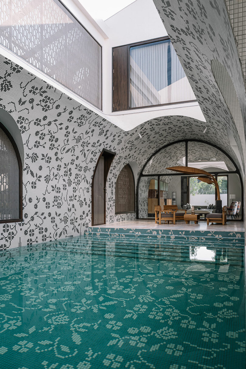 Indoor swimming pool with mosaic tile design on walls and ceiling, featuring arched doorways and large windows. Wooden furniture is visible in the lounge area adjacent to the pool.