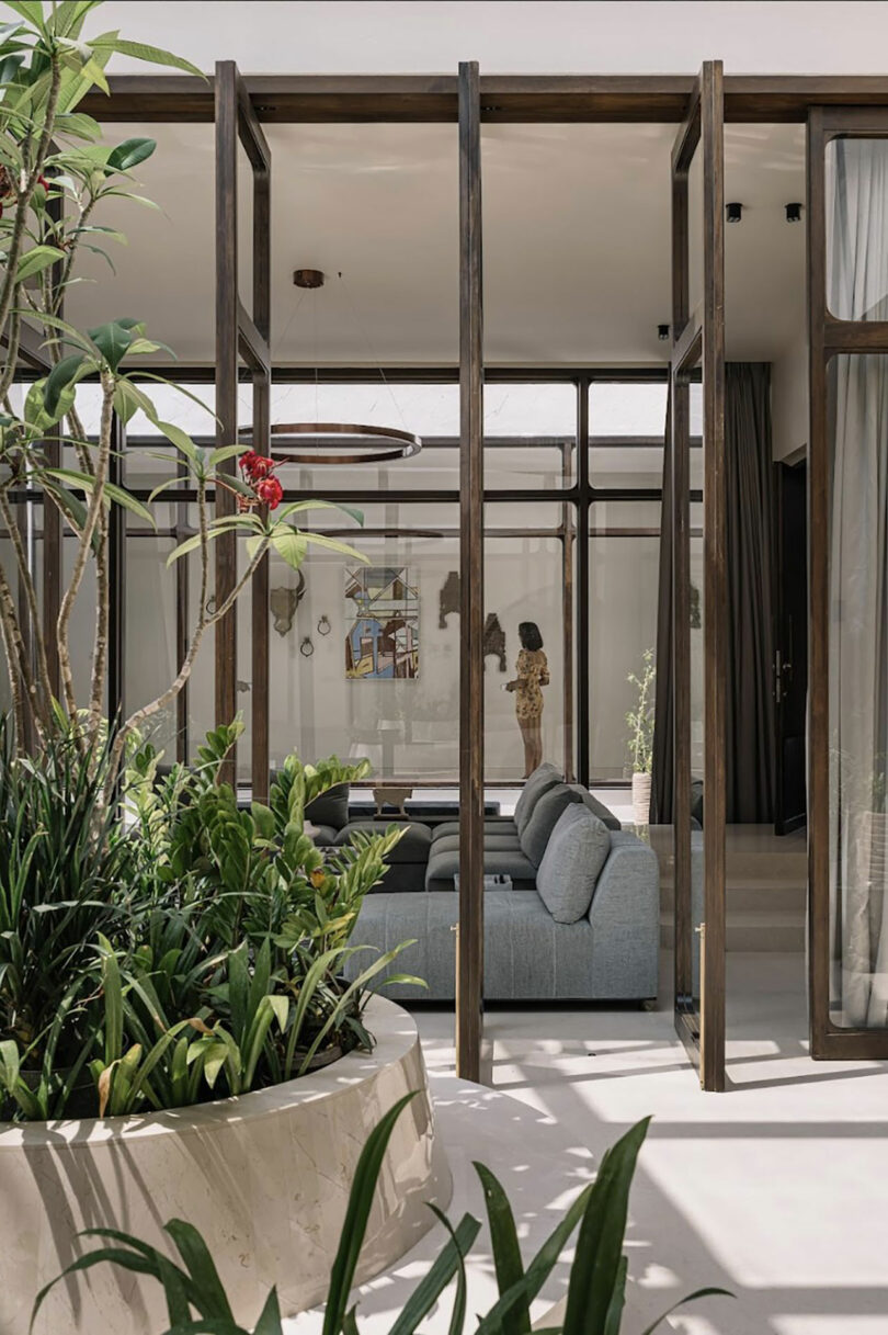 Modern indoor space with a mix of wooden beams, glass, and greenery. A gray couch is centered, with plants in the foreground and a person visible in the background. Artwork is hung on the far wall.