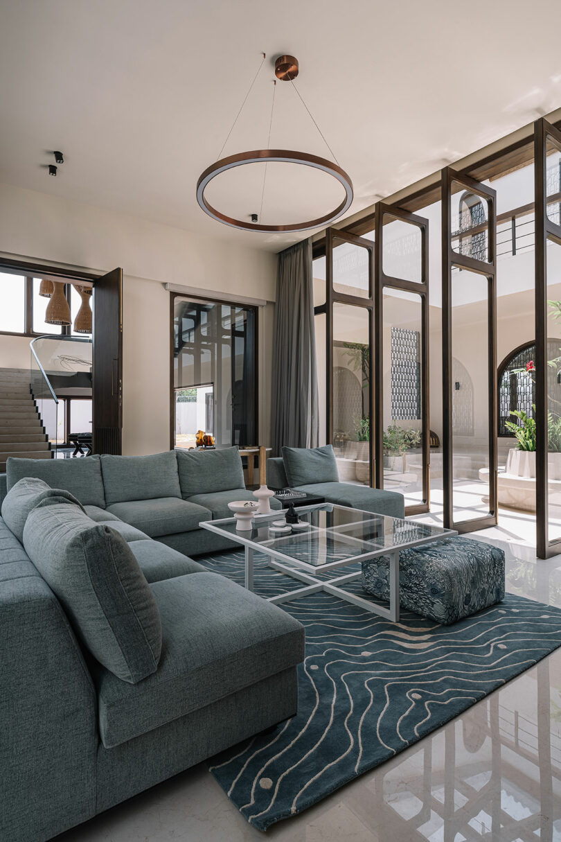 A modern living room with a large sectional sofa, a glass coffee table, and floor-to-ceiling windows. A circular light fixture hangs above, and a patterned rug covers the floor.