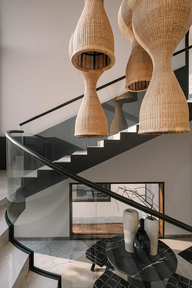 A modern staircase with glass railings curves upward. Woven pendant lights hang above. Below, a black table holds ceramic vases next to a window. Walls are white and floors are marble.