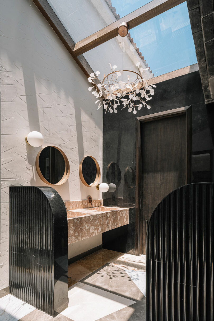 A bathroom with a skylight, featuring a marble sink, two round mirrors, wall-mounted round lights, and a decorative chandelier. The space has dark and white tiled walls and floors.