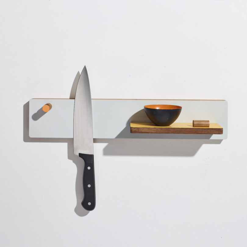 A knife, bowl, and wooden block rest on a minimalist wall-mounted shelf with a white and wooden design