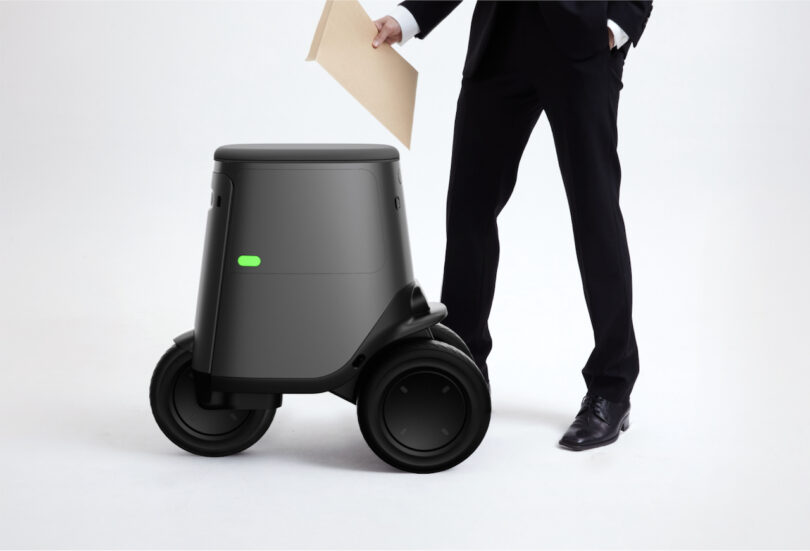 A person in a suit interacts with a small, autonomous delivery robot on wheels holding an envelope