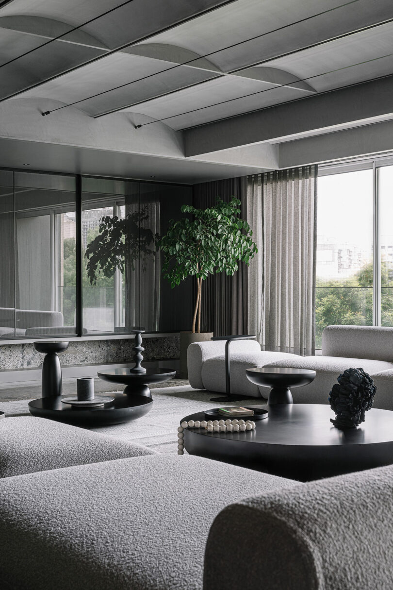 Modern living room with grey sofas, large windows, and a potted plant. Black decorative items sit on low tables, and curtains are partially drawn. Minimalist design with natural light.