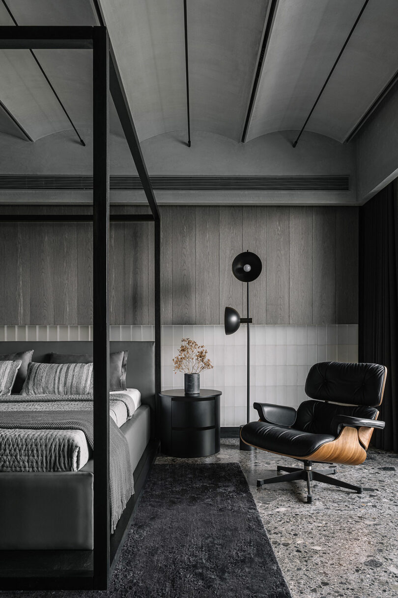 Modern bedroom with a black four-poster bed, matching side table, gray linens, a stylish black chair, wall-mounted lamps, and a gray-textured floor.