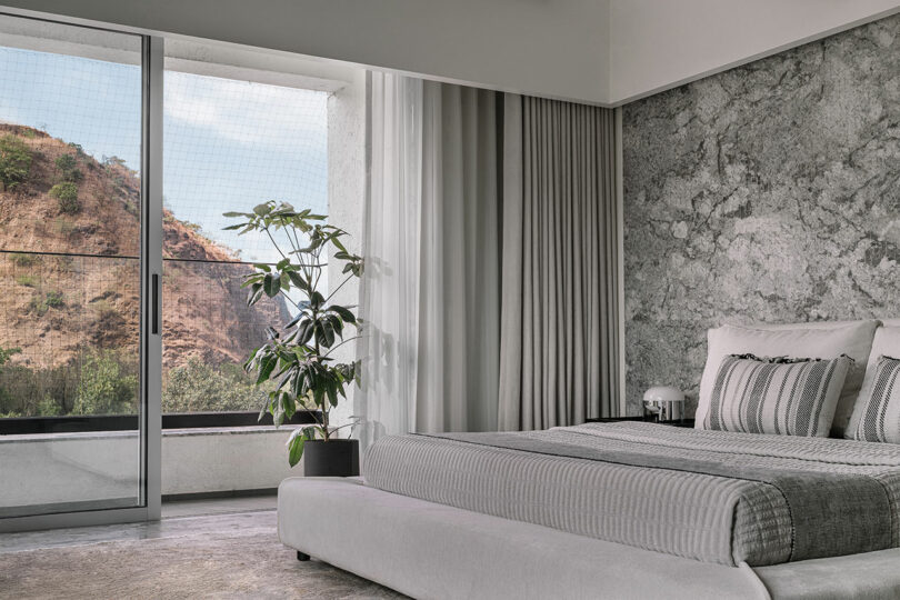 Minimalist bedroom with a large bed, gray textured wall, a potted plant, and a window with a view of a rocky landscape.