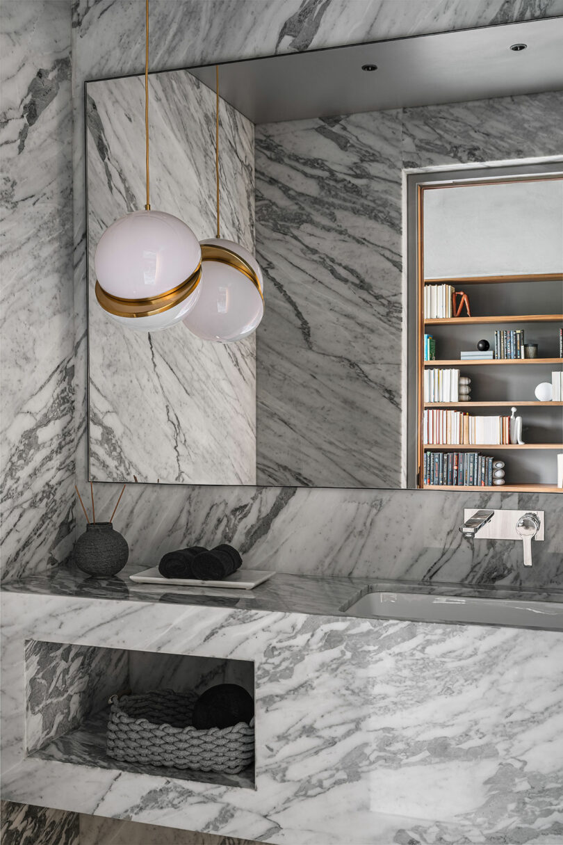 Bathroom with gray marble walls and countertop, a large mirror, a wall-mounted faucet, and two spherical pendant lights. A recessed shelf with books is visible in the reflection.