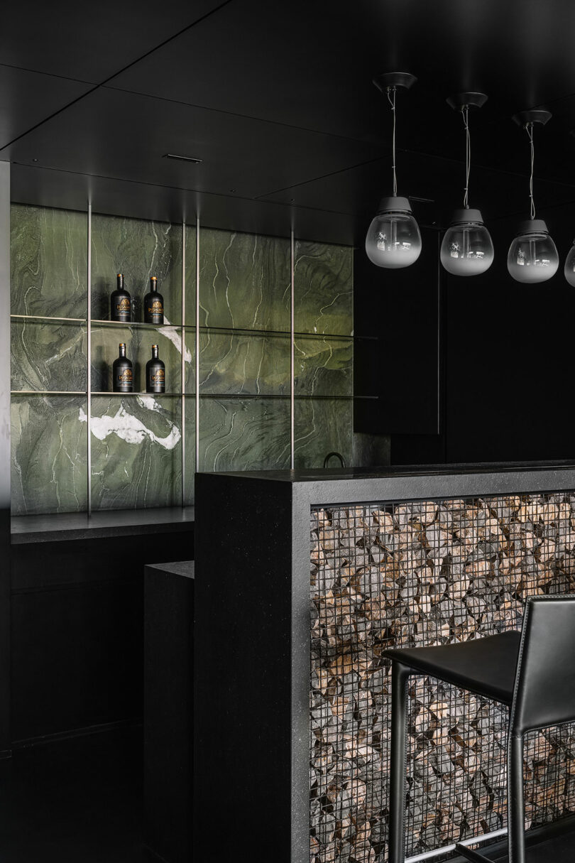 A modern bar area with a green wall backsplash, shelves with bottles, a stone-filled bar counter, and four hanging lights.