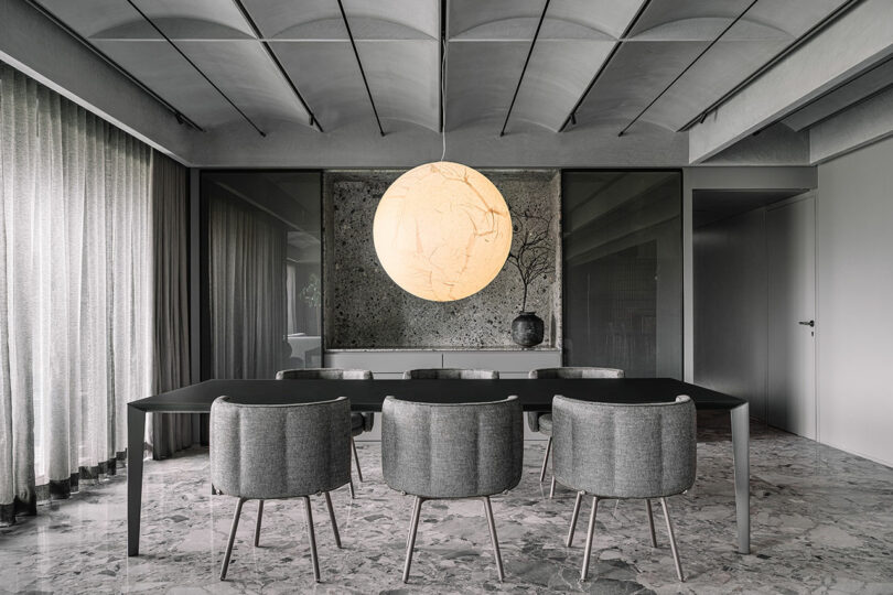 Modern dining room with a long table, six gray chairs, and a large, spherical light fixture. Curtains cover the windows, and a textured wall with a vase completes the minimalist design.