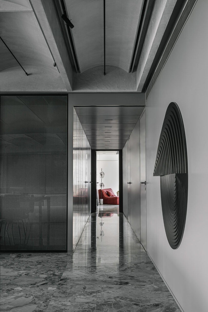 Modern hallway with sleek design, polished marble floor, and a large black circular wall art. A red chair is visible at the end of the hall, near a bright window.